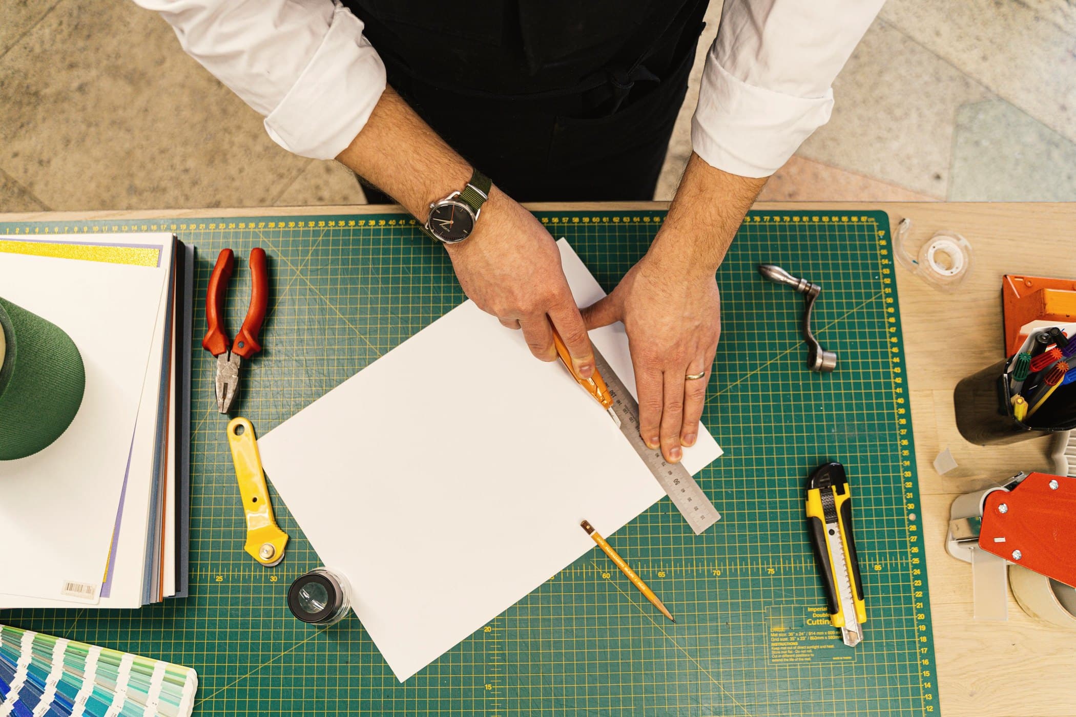 Overhead view of hands cutting a sheet of white board against a steel rule on a green cutting mat